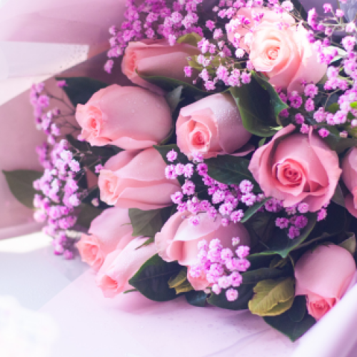 A bouquet of pink roses mixed with small purple flowers, wrapped in light paper, creating an elegant and charming floral arrangement.