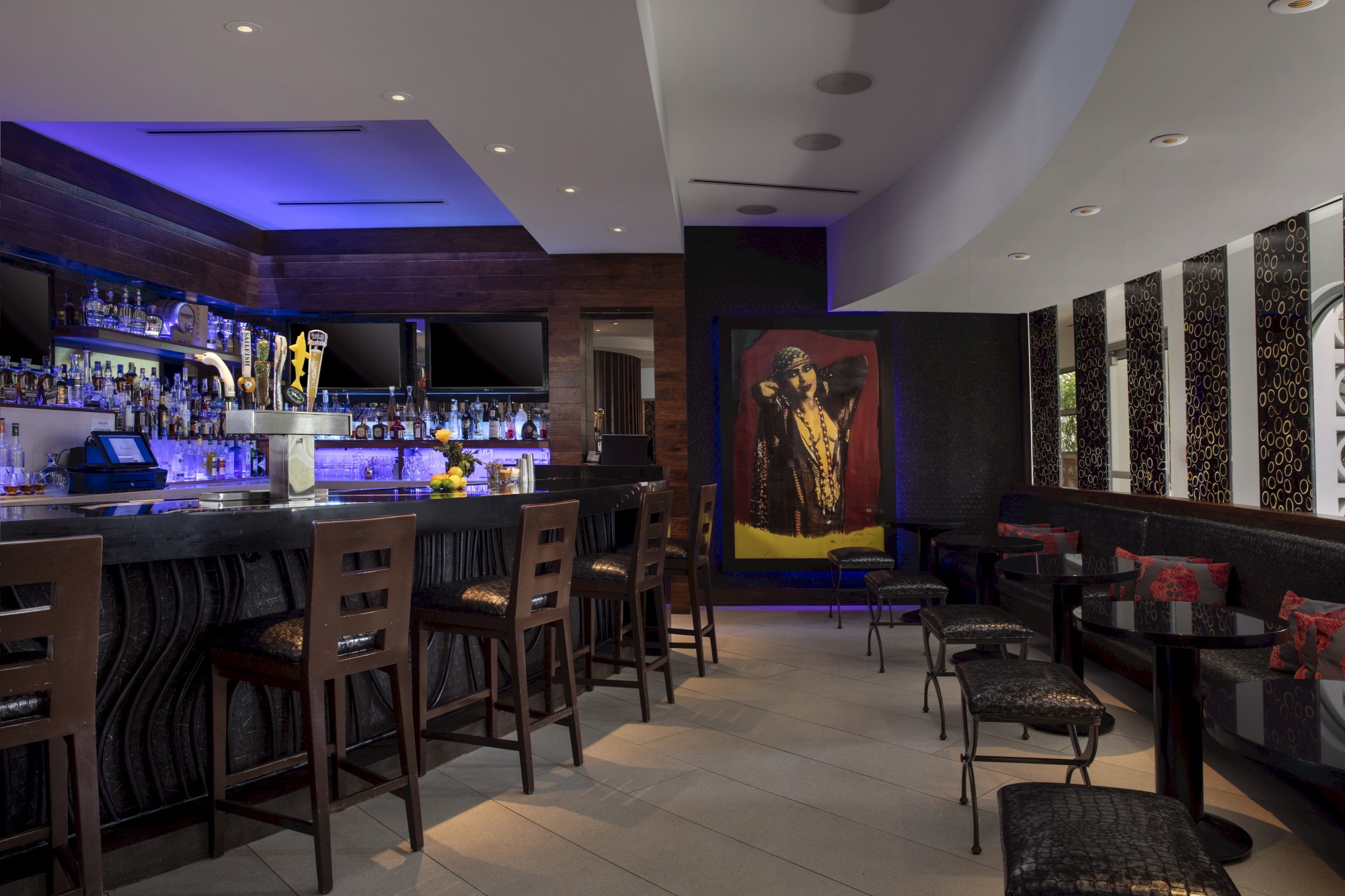 A stylish bar with high stools, a well-stocked counter, ambient lighting, and a colorful artwork on the wall, creating a cozy atmosphere.