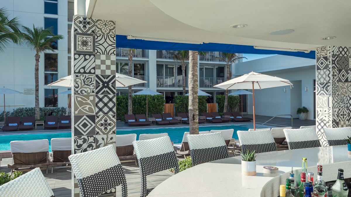 A poolside bar with patterned pillars, chairs, and a variety of bottles, set against a backdrop of lounge chairs and umbrellas.