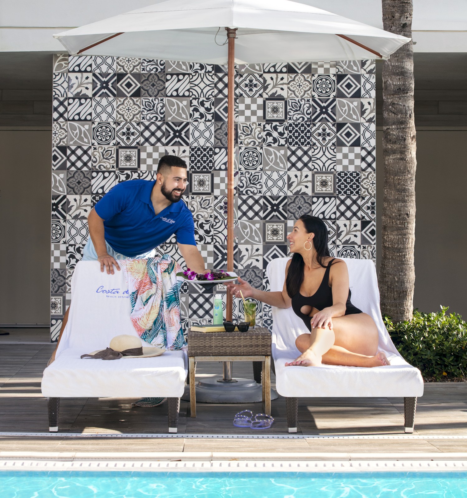 A man and woman relax by a pool on lounge chairs under an umbrella, against a patterned wall, with drinks and a table nearby.