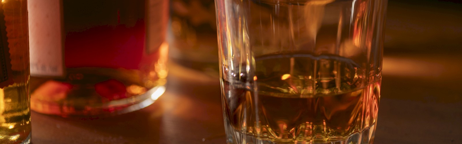 A glass filled with amber liquid, likely whiskey, is on a wooden surface, surrounded by bottles in warm lighting.