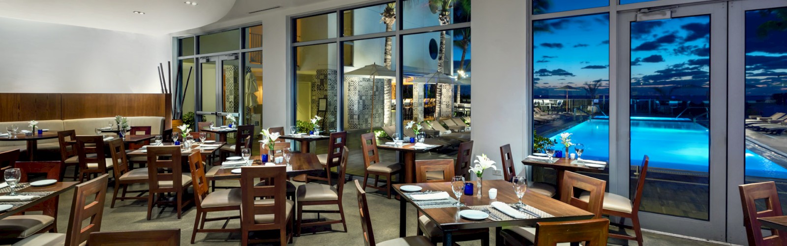 A modern restaurant with wooden tables and chairs, set for dining, overlooking a pool and an ocean view through large windows.