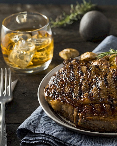 A grilled steak with roasted potatoes on a plate, served with a glass of whiskey. The setting includes a fork and rosemary for garnish.