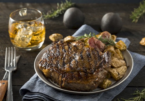 A grilled steak with roasted potatoes on a plate, served with a glass of whiskey. The setting includes a fork and rosemary for garnish.