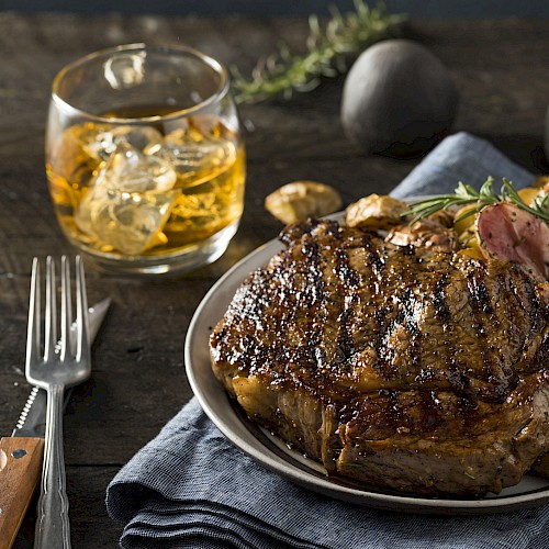 A grilled steak with roasted potatoes on a plate, served with a glass of whiskey. The setting includes a fork and rosemary for garnish.