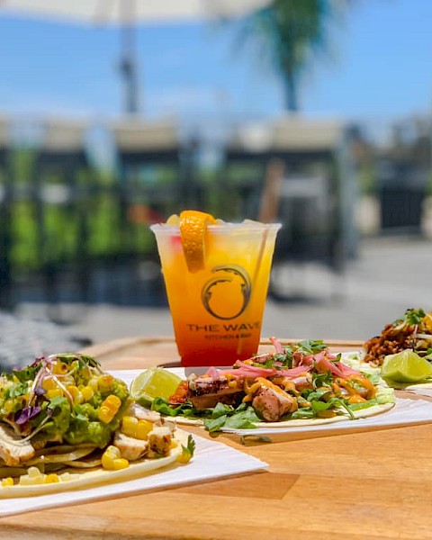 Three tacos with various toppings and a tropical cocktail are on a wooden table outdoors, with a blurred background of chairs and trees.