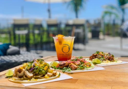 Three tacos with various toppings and a tropical cocktail are on a wooden table outdoors, with a blurred background of chairs and trees.
