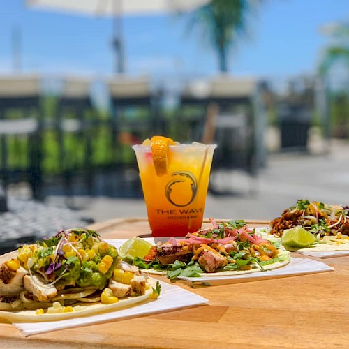 Three tacos with various toppings and a tropical cocktail are on a wooden table outdoors, with a blurred background of chairs and trees.