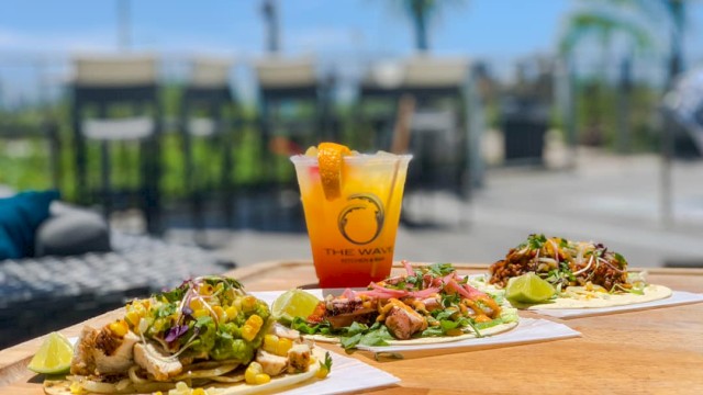 Three tacos with various toppings and a tropical cocktail are on a wooden table outdoors, with a blurred background of chairs and trees.