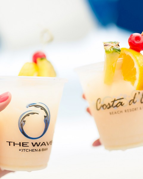Two hands holding fruity cocktails with garnishes in branded cups against a blurred background.