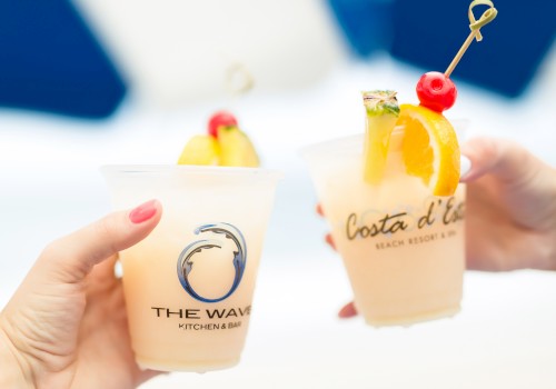 Two hands holding fruity cocktails with garnishes in branded cups against a blurred background.