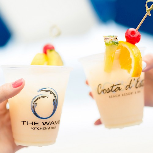 Two hands holding fruity cocktails with garnishes in branded cups against a blurred background.
