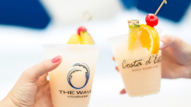 Two hands holding fruity cocktails with garnishes in branded cups against a blurred background.