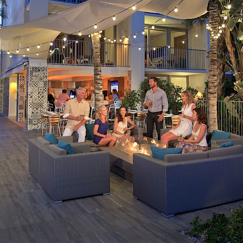 A group of people are relaxing around a fire pit on a patio with string lights and palm trees, enjoying an evening gathering.