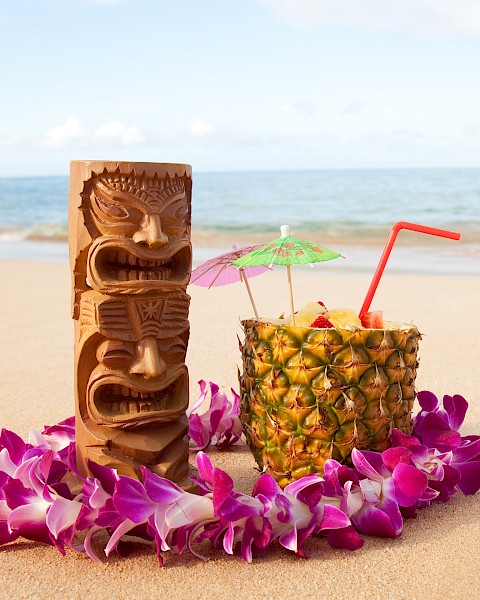 A tropical beach scene with a tiki statue, a pineapple drink with umbrellas, a straw, and a lei of purple flowers on the sand.