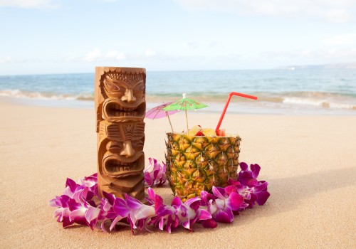 A tropical beach scene with a tiki statue, a pineapple drink with umbrellas, a straw, and a lei of purple flowers on the sand.