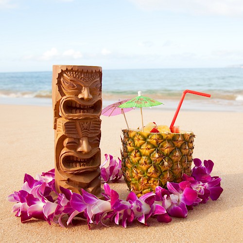 A tropical beach scene with a tiki statue, a pineapple drink with umbrellas, a straw, and a lei of purple flowers on the sand.