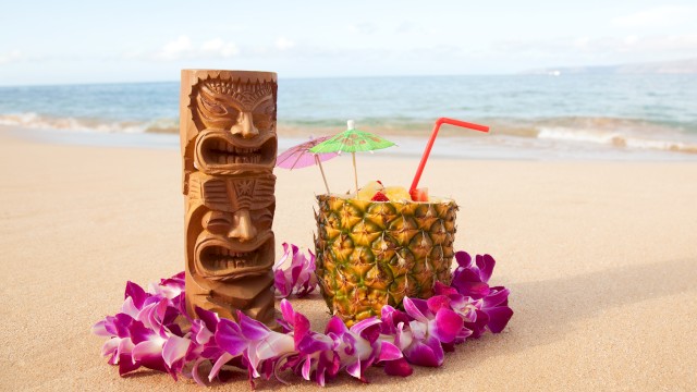 A tropical beach scene with a tiki statue, a pineapple drink with umbrellas, a straw, and a lei of purple flowers on the sand.