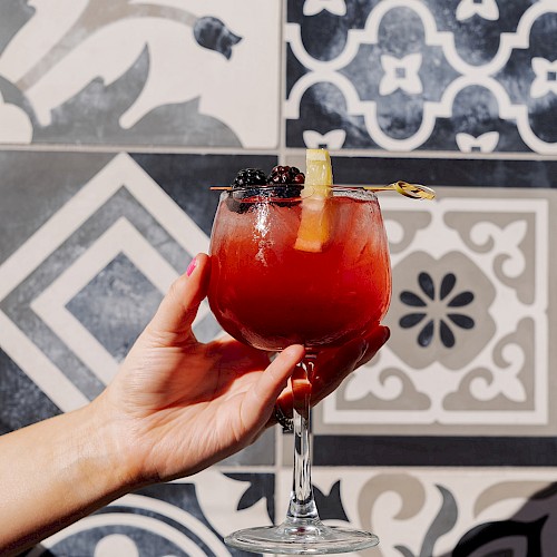 A hand holds a glass with a red cocktail, garnished with fruit, against a backdrop of decorative black and white tiles.