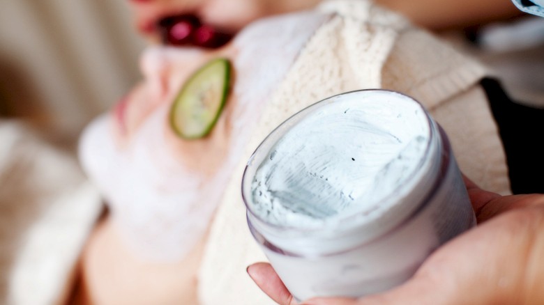 A person is receiving a spa treatment, with a facial mask applied and a cucumber slice on one eye, while holding a jar of cream.