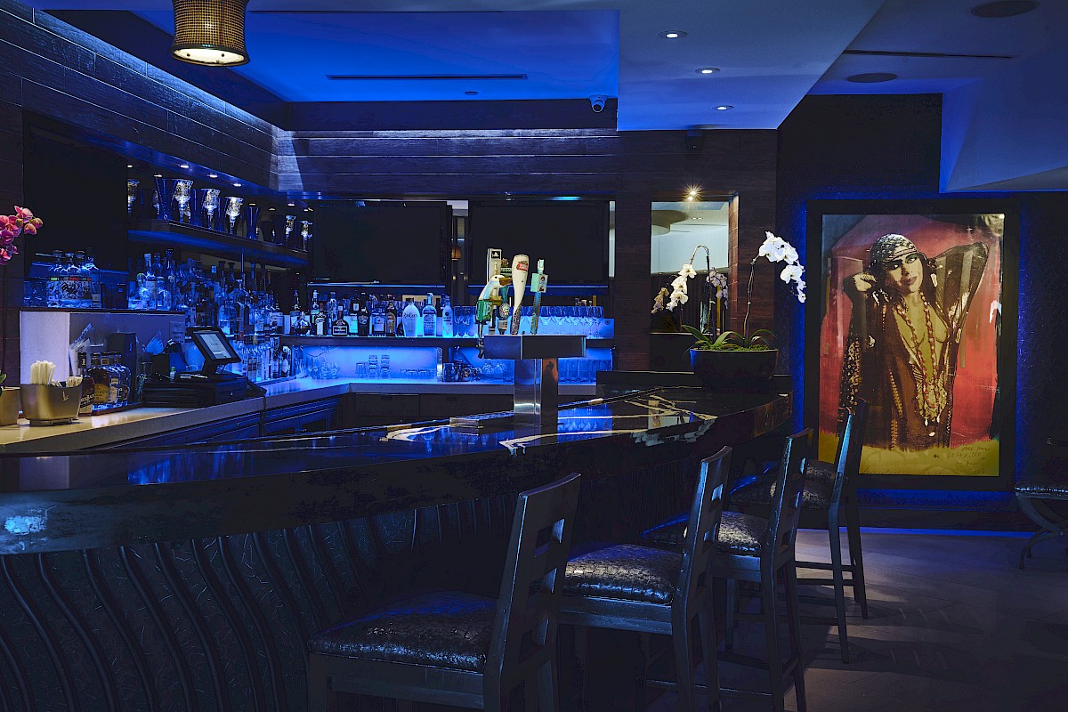 A dimly lit bar features a blue glow, with high chairs, a wide selection of drinks, and decorative artwork near the entrance.