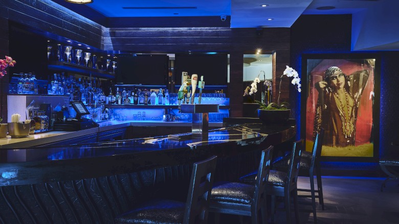 A dimly lit bar features a blue glow, with high chairs, a wide selection of drinks, and decorative artwork near the entrance.