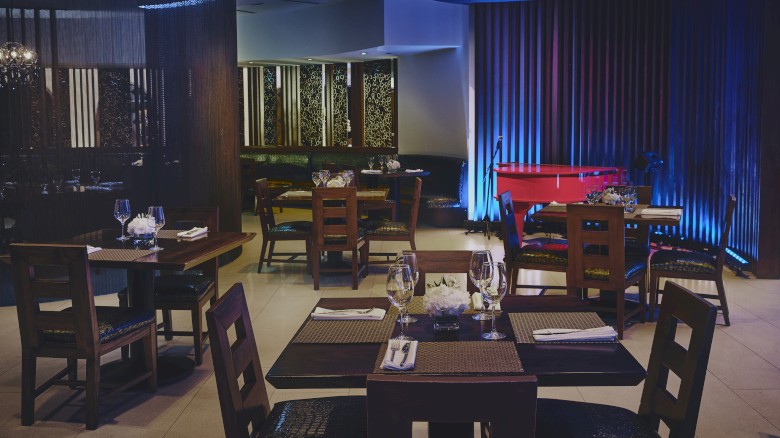 A dimly lit restaurant with wooden tables, chairs, place settings, and ambient blue lighting, creating a cozy atmosphere.