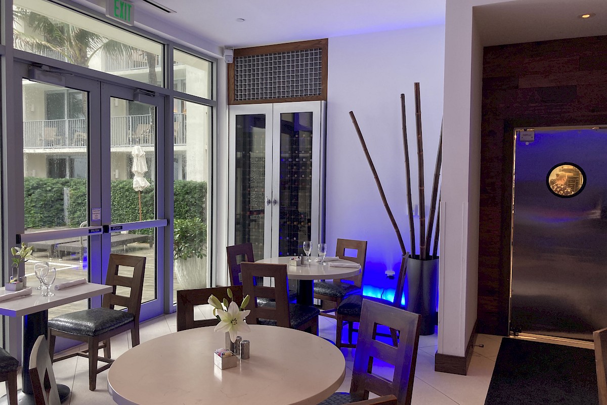 A modern, cozy restaurant interior with round tables, chairs, large windows, and stylish decor including tall vases and ambient lighting.