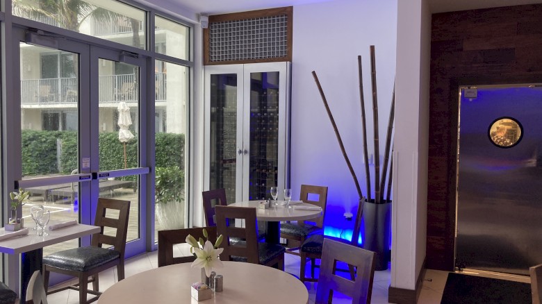 A modern, cozy restaurant interior with round tables, chairs, large windows, and stylish decor including tall vases and ambient lighting.