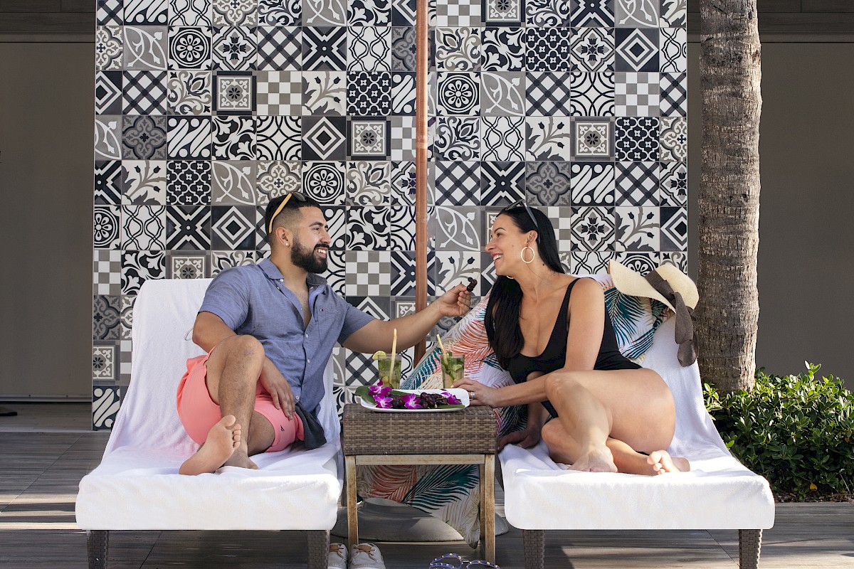 A couple sits on lounge chairs by a pool under an umbrella, surrounded by stylish tiles and palm trees.