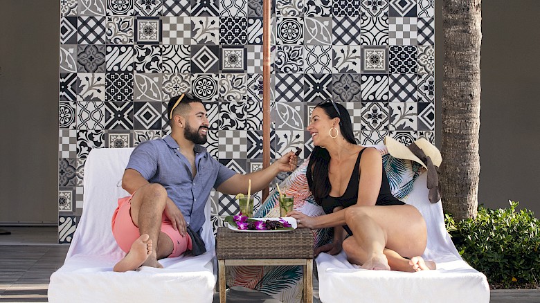 A couple sits on lounge chairs by a pool under an umbrella, surrounded by stylish tiles and palm trees.