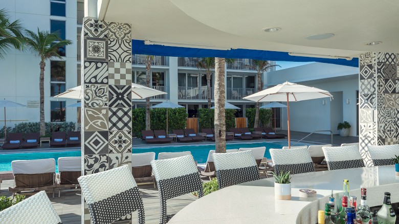 Outdoor bar with patterned pillars, wicker stools, a display of bottles, and a view of a pool with sun loungers and umbrellas.