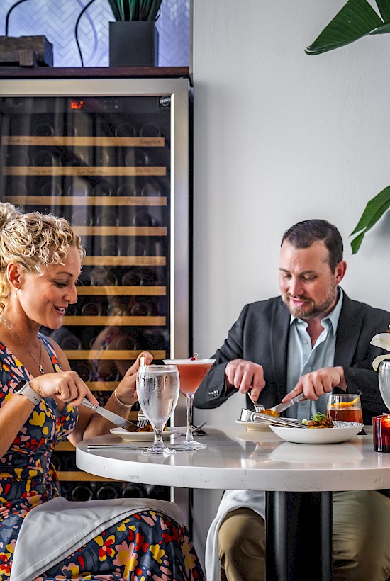 A couple is dining at a restaurant table with drinks and meals, surrounded by plants and a wine cellar in the background.