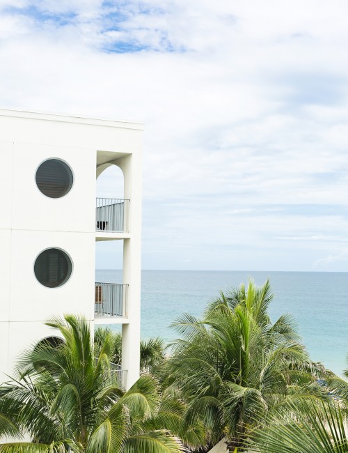 A white building with circular windows is on the left, overlooking palm trees and a blue ocean under a cloudy sky.