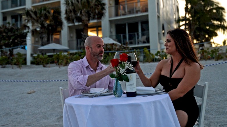 A couple is enjoying a romantic dinner at a table set with wine and roses on a beach, in front of a building at sunset.