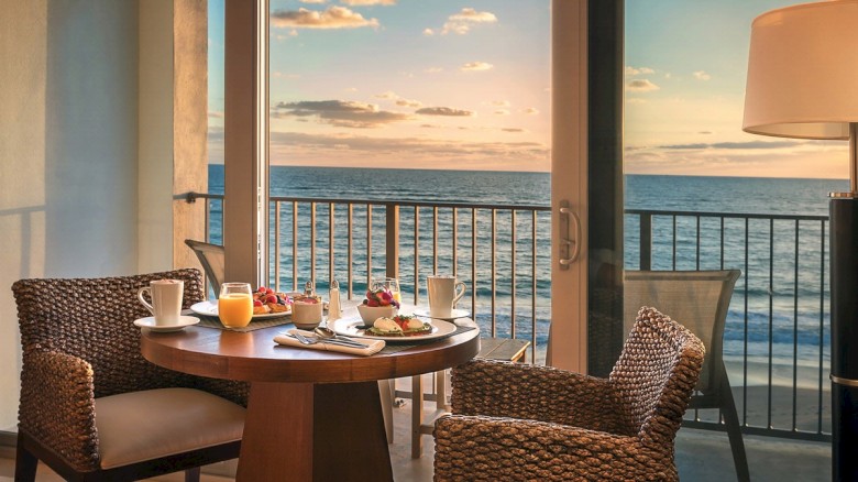 A breakfast setup on a table by a window overlooking the ocean, featuring juice, coffee, fruit, and pastries in a cozy setting.