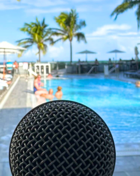 A microphone is in the foreground with a poolside setting featuring lounge chairs, people sunbathing, and palm trees in the background.