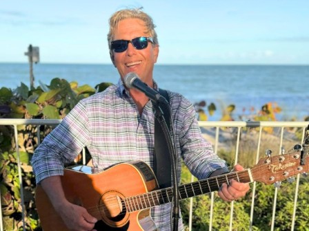 A person is playing an acoustic guitar and singing outdoors by the sea, wearing sunglasses and a plaid shirt, with a microphone stand.