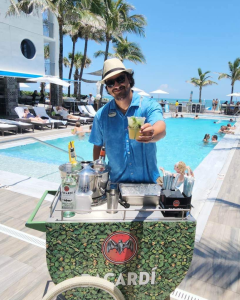 A person in a hat serves drinks by a pool with palm trees and people lounging under umbrellas in the background.
