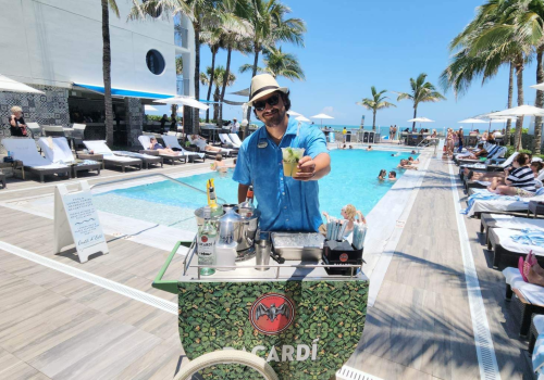 A person in a hat serves drinks by a pool with palm trees and people lounging under umbrellas in the background.