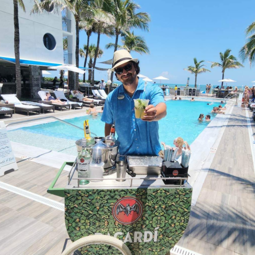 A person in a hat serves drinks by a pool with palm trees and people lounging under umbrellas in the background.
