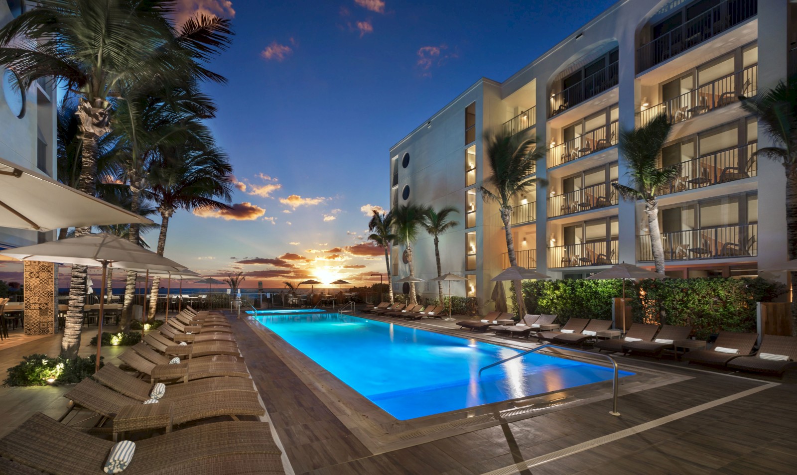 The image shows a luxurious hotel pool area at sunset, with lounge chairs, palm trees, and a modern building in the background.