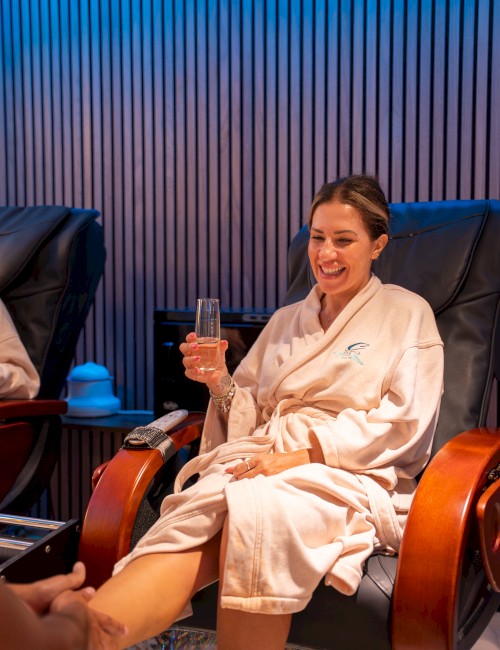 A person in a spa robe enjoys a foot massage while holding a glass, seated in a reclining chair with a relaxed expression.