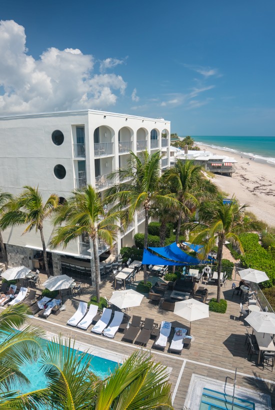 A beachfront hotel with a pool, surrounded by palm trees, sun loungers, umbrellas, and ocean views on a sunny day.