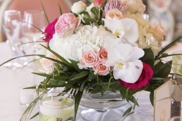 A floral arrangement with pink, white, and green flowers on a table, surrounded by candles and glassware in a decorative setting.