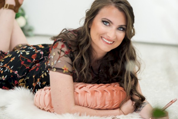 A person is lying on a rug with a pillow, writing in a notebook, and surrounded by flowers, wearing a floral dress and heeled sandals.