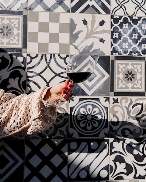 A hand holding a wine glass is in front of a wall with assorted black and white patterned tiles, creating a stylish backdrop.