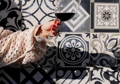 A hand holding a wine glass is in front of a wall with assorted black and white patterned tiles, creating a stylish backdrop.