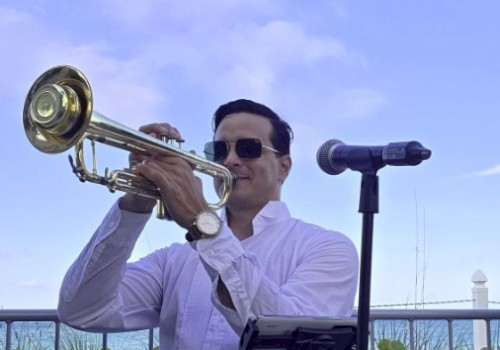 A man with sunglasses plays a trumpet outdoors, standing in front of a microphone with a scenic sky and greenery in the background.