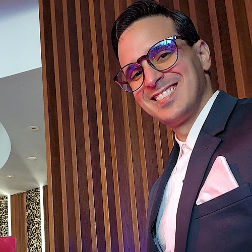 The image shows a person in a suit with glasses, smiling in a stylish setting with wood paneling and leopard print details on the walls.